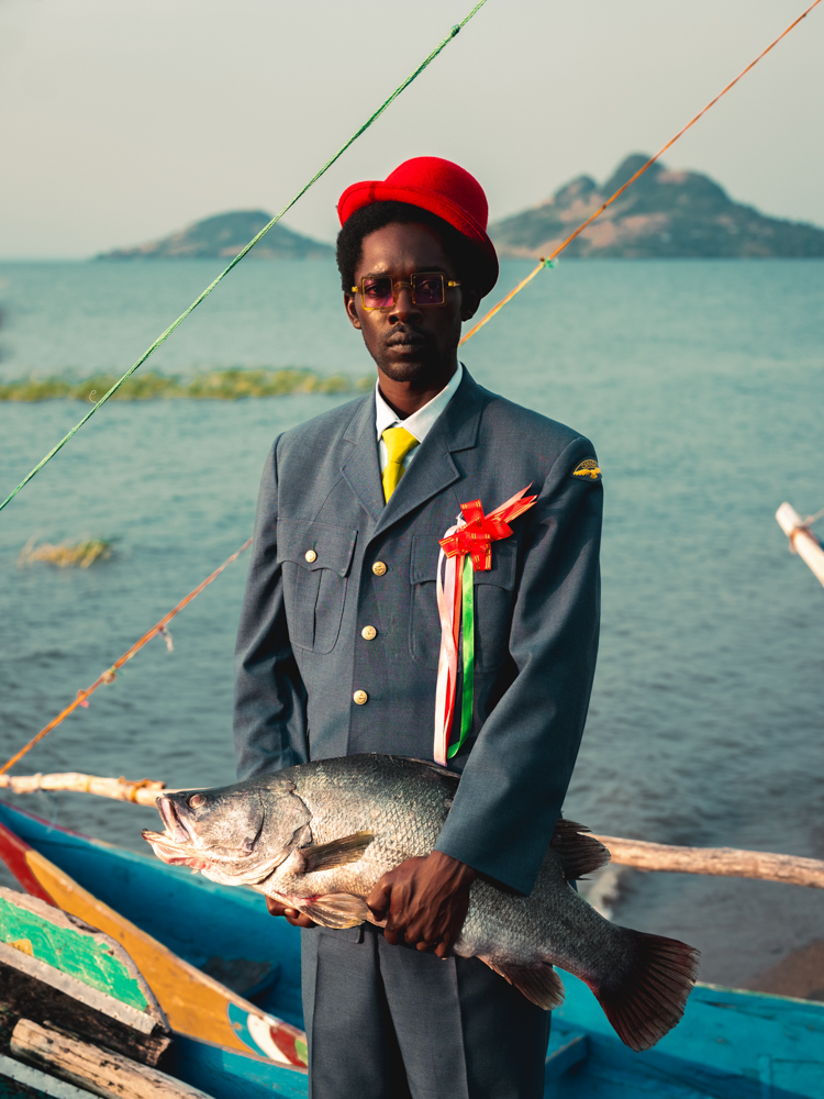 a man in a suit and a red hat, holding a large fish in his hands, standing infront of lake victoria.