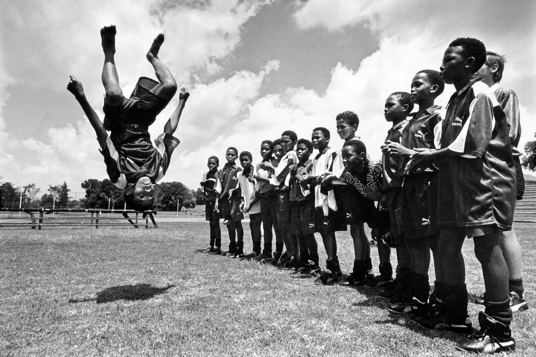 Steven Pienaar performing a backflip on a football field as children cheer him on in the background.