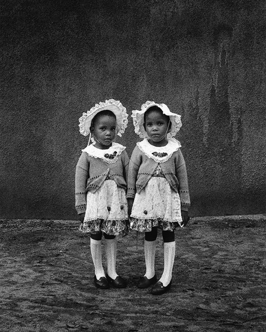 black and white photo of two twins, standing posing for the camera. Photo taken by Jodi Bieber